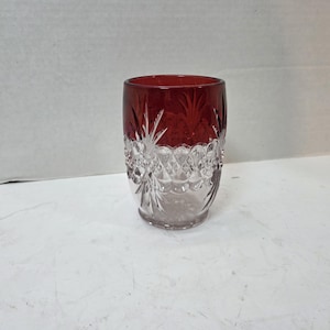 Puede incluir: Un vaso cilíndrico que presenta una sección superior de color rojo intenso y una sección inferior transparente. La superficie exterior está decorada con intrincados patrones de vidrio tallado, incluyendo motivos de estrellas.