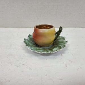 Könnte beinhalten: Ein Keramik-Teebecher- und Untertassen-Set. Der Teebecher ist wie ein Pfirsich geformt, mit einem Pfirsich- und Gelbverlauf und einem grünen Griff. Die Untertasse ist blattförmig und grün. Das Innere der Tasse ist goldfarben.