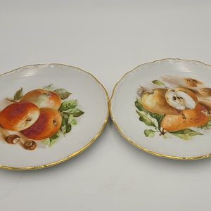 May include: Two white porcelain plates with gold trim, each featuring a different fruit design. One plate has three apples, while the other has three pears.
