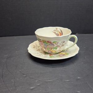May include: A cream-colored teacup and saucer set with a floral design featuring purple, yellow, and red accents. The cup has a scalloped rim and a green handle. The saucer is round with a matching floral pattern.