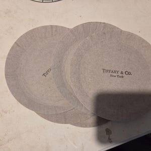 Peut inclure: Plusieurs dessous de verre circulaires gris en papier, avec le logo "Tiffany & Co. New York" imprimé. Les dessous de verre se chevauchent légèrement, suggérant qu'ils font partie d'un ensemble.