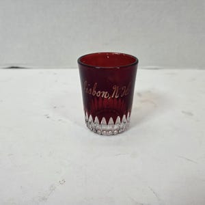 Peut inclure: Un petit verre à liqueur en verre rouge profond doté d'une base transparente à facettes avec un motif en dents de scie. L'extérieur du verre est gravé avec le texte cursif Lisbon, NH, apparaissant en écriture claire sur le verre rouge foncé.