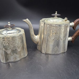 Könnte beinhalten: Ein silbernes Teekannen- und Zuckerdose-Set mit einem verzierten Design. Die Teekanne hat einen Holzgriff und einen silbernen Deckel mit einem Knopf. Die Zuckerdose hat einen silbernen Deckel mit einem Knopf.