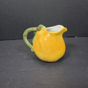May include: A ceramic pitcher shaped like a lemon, with a textured yellow body and a green handle. The pitcher has a white interior and a spout for pouring. The handle is green with a leaf detail.