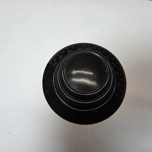 Puede incluir: Un dial circular negro con una serie de números y marcas alrededor del borde. El dial tiene un centro elevado y escalonado. Los números parecen estar en incrementos de diez, con marcas para cada unidad individual. El dial está sobre un fondo blanco.