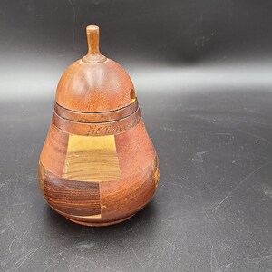 Peut inclure: Un récipient en bois en forme de poire avec un couvercle, fabriqué à partir de différents types de bois, avec une palette de couleurs brun foncé, brun rougeâtre et brun clair.