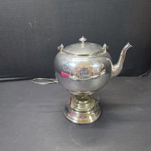 Puede incluir: Una tetera de metal plateado con cuerpo redondeado, pico curvo y tapa con un pequeño pomo. La tetera está montada sobre un soporte circular de metal a juego que cuenta con un quemador central y un asa lateral para facilitar su transporte.