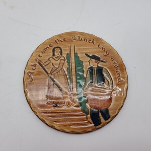 May include: A round, wooden plaque with a painted scene of a woman sweeping and a man carrying a bucket. The plaque has the text "Alch' come the back way around" written around the edge.