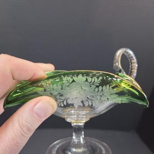 Op de afbeelding: Een groene glazen serveerschaal met een handvat. De schaal heeft een bloemenontwerp dat in het glas is ge&euml;tst. De rand is afgewerkt met goud. De schaal wordt door een hand vastgehouden.