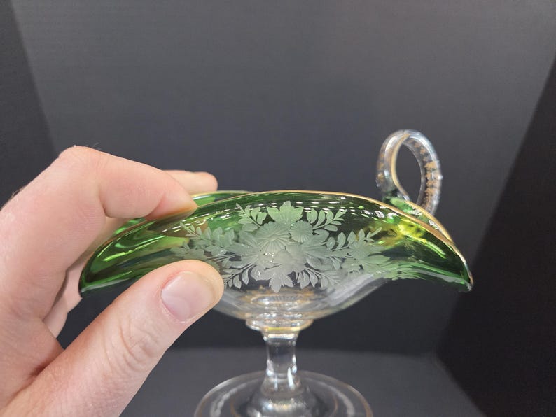 Op de afbeelding: Een groene glazen serveerschaal met een handvat. De schaal heeft een bloemenontwerp dat in het glas is ge&euml;tst. De rand is afgewerkt met goud. De schaal wordt door een hand vastgehouden.