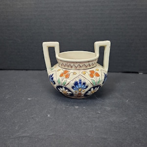 May include: A small, cream-coloured ceramic vase with two handles. The vase is decorated with a floral pattern in blue, orange, and green. The rim has a brown zigzag design.