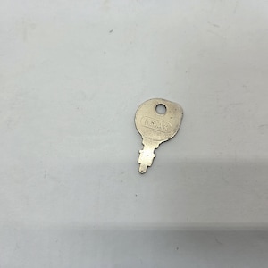 May include: A silver-colored key with the text "INDAK" stamped on the head. The key has a circular hole at the top and a notched blade. The key is on a white surface.