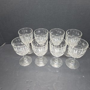 May include: A collection of eight clear, stemmed glasses. Each glass features a decorative etched pattern on the bowl and a delicate floral design. The glasses are arranged on a dark surface.