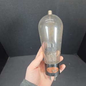 May include: A vintage vacuum tube, featuring a clear glass bulb and a black base. The tube has a cylindrical form, with internal components visible through the glass. The base has several pins and an orange and black label.