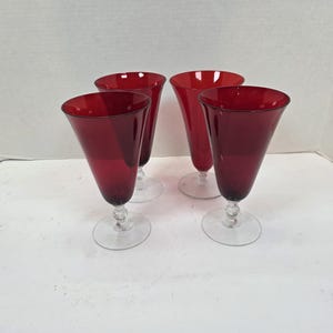May include: Four ruby red wine glasses with a flared, conical bowl and a clear, decorative stem. The glasses are set on a clear, circular base. The glasses are empty and are arranged on a white surface.