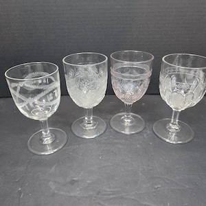 Pode incluir: Quatro taças de vinho transparentes com diferentes desenhos gravados. Cada taça apresenta um padrão único, incluindo motivos florais e geométricos. As taças estão dispostas em fila sobre uma superfície escura.