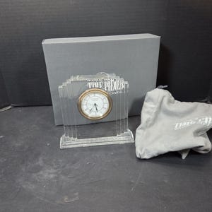 May include: A clear, Art Deco-style clock with a gold-tone bezel and white face. The clock is in front of a gray box and a gray cloth bag with the words "Time Pieces" printed on it.