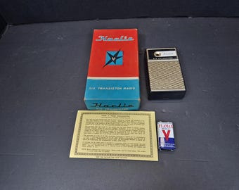Może przedstawiać: Zestaw radia Harlie Six Transistor w stylu vintage, zawierający oryginalne pudełko, radio, gwarancję i baterię Flamax. Pudełko jest czerwono-niebieskie z logo Harlie. Radio jest czarno-srebrne.