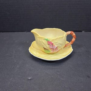 以下が含まれることがあります： 小さな黄色のクリーマーとソーサーのセット。クリーマーは葉のような質感で、赤と茶色のストライプのハンドルが付いています。セットはピンク、緑、黄色の花柄で装飾されています。どんなティーセットにも魅力的な追加です。