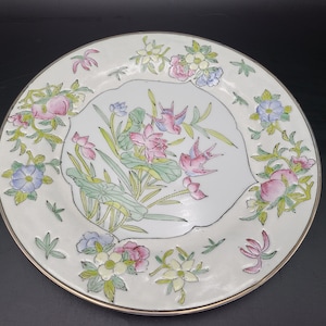 May include: A white ceramic plate with a gold rim features a floral design with pink, blue, and green flowers and birds. The plate has a raised rim and a scalloped edge.