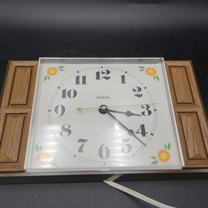 May include: A brown and white electric wall clock with a wood-grain frame and floral accents. The clock face features black Roman numerals and a white background. The clock is plugged in with a white cord.
