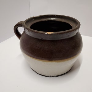 Könnte beinhalten: Ein brauner und weißer glasierter Keramiktopf mit einem einzigen Henkel. Der Topf hat eine breite Öffnung und einen runden Körper.
