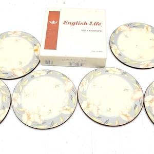 May include: A set of six coasters featuring floral patterns. Each coaster has a cream-colored center with a floral design in pink, yellow, and green, framed by a grey border. The coasters are packaged in a white box with the text "English Life, six coasters."