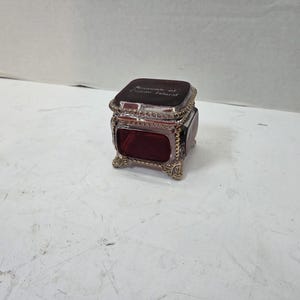 May include: A small, square, deep red glass trinket box with a hinged lid. The box features ornate, metallic gold-toned filigree trim along the edges and four decorative feet. The lid is engraved with the text "Souvenirs of Coney Island".