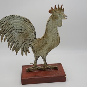 May include: A white painted metal rooster figurine with a red wooden base. The rooster has a raised tail and a detailed head with a comb and wattle.
