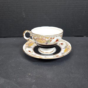 May include: A vintage teacup and saucer set. The white teacup has a gold handle and rim, with a decorative floral pattern. The saucer has a matching design with a black and gold centre. A classic tea set.