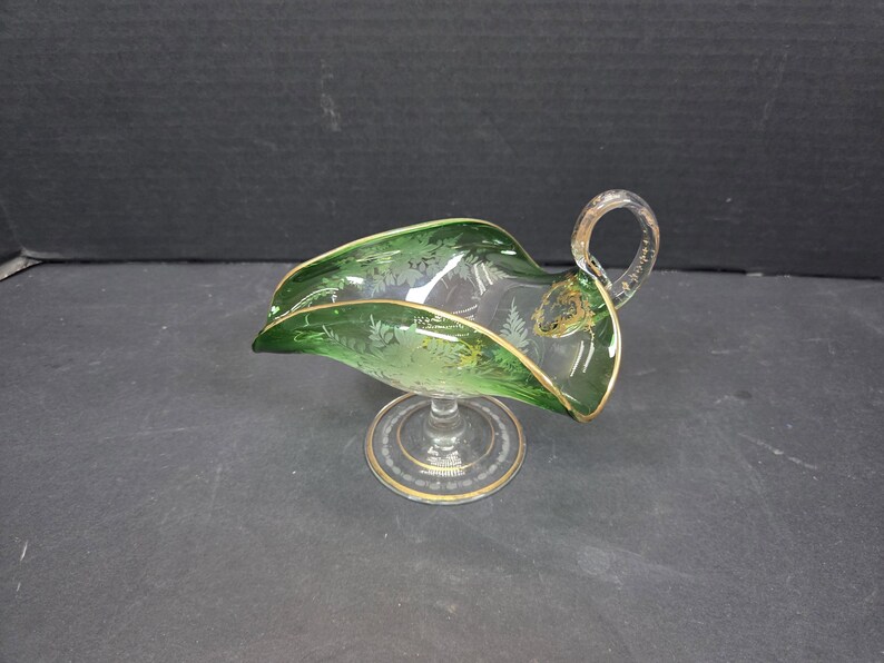 Op de afbeelding: Een heldere glazen juskom met een groene rand en handvat, geaccentueerd met gouden rand. De kom heeft een voetstuk en ge&euml;tste bloemmotieven. Een decoratief serveerstuk.