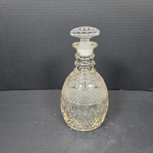 May include: A clear glass decanter with a decorative stopper. The decanter has a rounded base and a textured, diamond-patterned design. The stopper is a flat, circular shape with a starburst pattern. The decanter is approximately 8 inches tall.