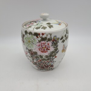 May include: White porcelain jar with a lid, decorated with a floral pattern of pink, green, and white flowers, and butterflies. The jar has a gold trim around the lid.