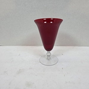 May include: A tall, ruby red glass goblet with a flared rim. The stem features a decorative, clear, beaded design, and the base is a clear, circular disc. The goblet is empty and sits on a white surface.