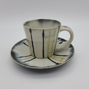 May include: A white ceramic teacup and saucer set with black and gold abstract designs.