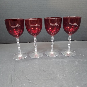 Puede incluir: Cuatro copas de vino tinto rubí con tallos transparentes y perlados y bases redondas. Las copas están dispuestas en fila, mostrando su elegante diseño. Las copas son de cristal.