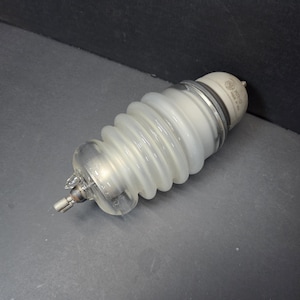 May include: A clear and white glass electrical insulator with a ribbed, accordion-like body. The insulator has a metal base and a white ceramic top with text. The object is on a grey surface.