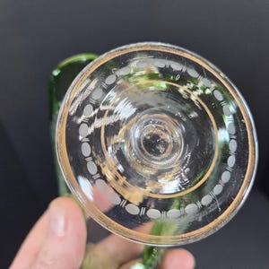Op de afbeelding: Helder glazen wijnglas met een gouden rand en decoratieve witte ovale etsen. De steel van het glas is groen. Het glas wordt door een hand tegen een donkere achtergrond gehouden.