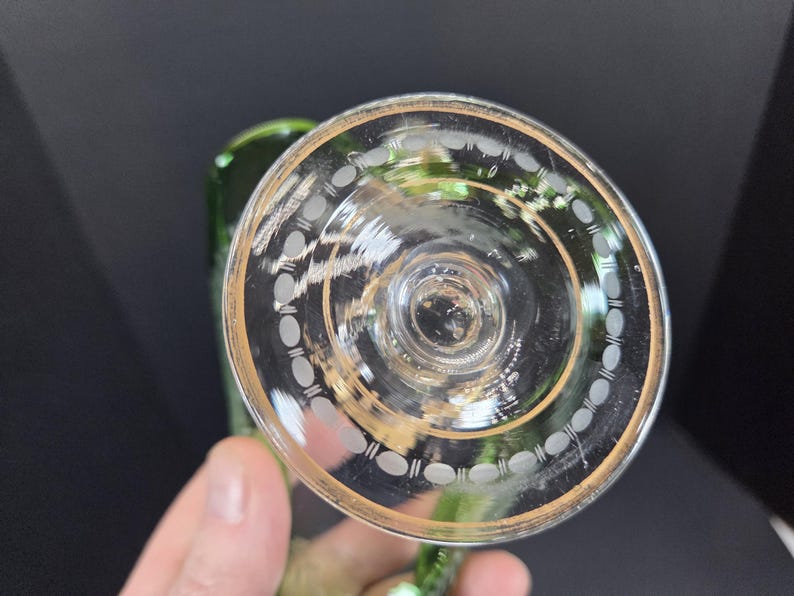 Op de afbeelding: Helder glazen wijnglas met een gouden rand en decoratieve witte ovale etsen. De steel van het glas is groen. Het glas wordt door een hand tegen een donkere achtergrond gehouden.