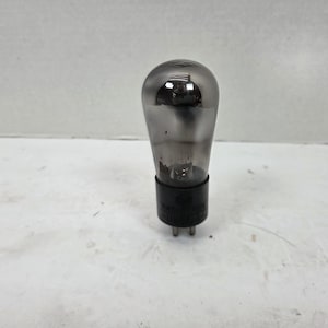 May include: A vintage vacuum tube, featuring a clear glass bulb and a black base. The tube's internal metallic structure is visible. The base has three pins for connection. The tube is likely used in electronic devices.