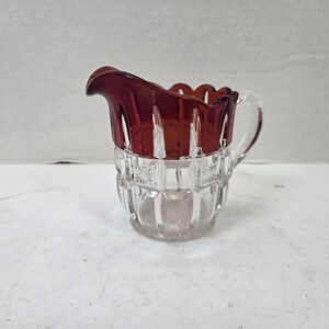 May include: A clear glass creamer pitcher featuring a deep red stained upper rim with a scalloped edge. The body of the pitcher displays a pressed geometric pattern with vertical panels and small starburst motifs, complemented by a curved handle.
