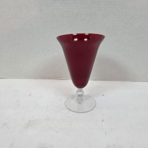 May include: A tall, ruby-red glass with a flared top and a clear, round base. The stem features a decorative, textured design. The glass is likely used for serving beverages.