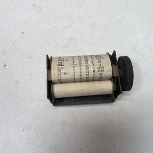Puede incluir: Un dispositivo antiguo de metal negro con dos rollos de papel blanco. El rollo superior muestra números y letras impresos. Un dial negro está en el lado derecho. Probablemente sea un registrador de datos antiguo o similar.