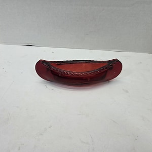 May include: A translucent red glass dish shaped like a canoe. The vessel features a smooth, curved exterior and a textured, rope-like pattern along the top rim. The glass is deep red in color, with a hollow center and pointed ends.