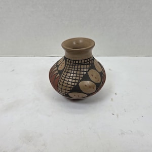 Puede incluir: Un pequeño jarrón redondo de cerámica con borde y cuerpo de color beige. El jarrón está decorado con un patrón geométrico de formas negras, rojas y beige. El diseño incluye un patrón de cuadrícula y formas circulares.