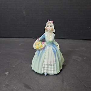 May include: A porcelain figurine of a woman in a long dress, holding a basket of flowers. The dress is light blue and green, with a white apron. The woman wears a white bonnet with a pink bow.