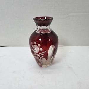May include: A small, bulbous glass vase featuring a deep ruby red color with clear, cut-glass patterns. The design includes etched circular motifs and geometric cross-hatching near the base, with a flared rim and a smooth, transparent neck.