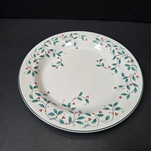 May include: White ceramic serving plate with a green and red holly berry design around the rim. The plate has a dark green border and is set against a black background. A festive piece for holiday gatherings.
