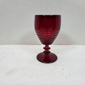 May include: A ruby red wine glass with a round base and a stem featuring a series of stacked, rounded elements. The bowl of the glass has a series of horizontal ridges. The glass is made of a transparent material.