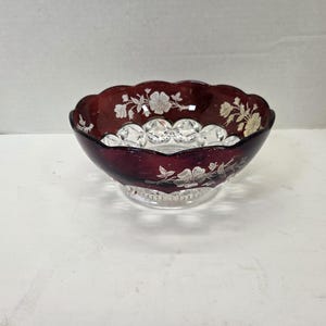 May include: A deep red glass bowl featuring a scalloped rim and a clear glass base with a faceted pattern. The exterior of the bowl is decorated with a silver-colored floral design that wraps around the upper portion of the dark red glass.